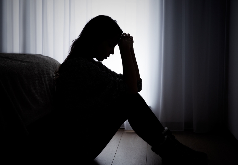 A woman in sillhouette, with her hands pulled to her forehead, sitting on a floor in frotn of a white curtain.